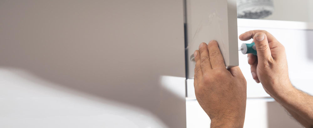 Close-up of a person's hands using a screwdriver to remove or install a white cabinet door handle on a white wall or cabinet.