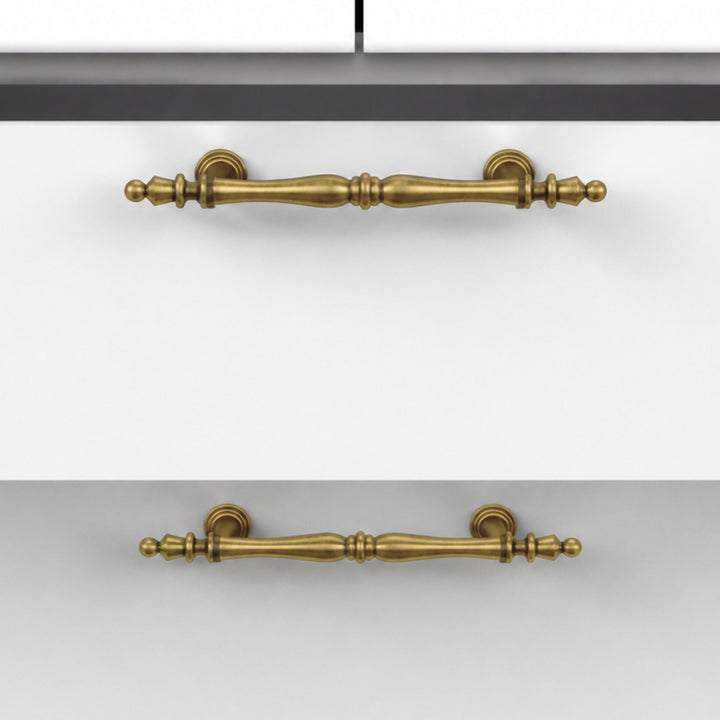 Two antique brass cabinet handles shown in a split view, mounted on a white cabinet door in the top half and a gray cabinet door in the bottom half.
