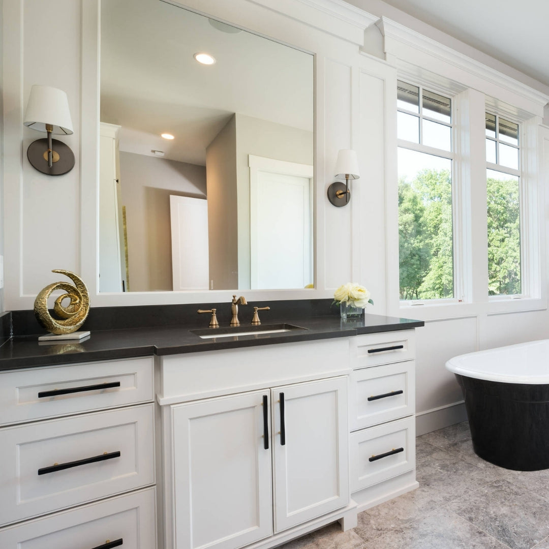 A white double-sink bathroom vanity with a black countertop, brass faucet, and a large wall mirror, shown in a bright, modern bathroom with windows, wall sconces, and a freestanding bathtub.